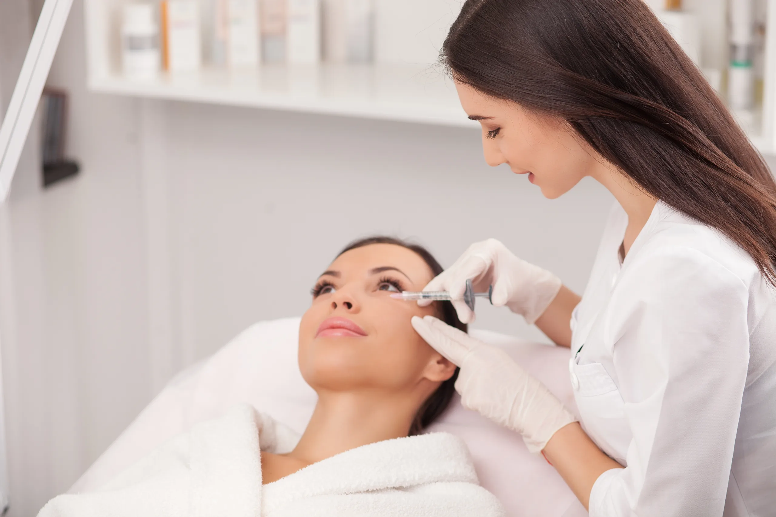 Attractive young woman is getting a collage injection in her face. She is sitting calmly at clinic. The expert beautician is filling female nasolabial wrinkles by hyaluronic acid and smiling